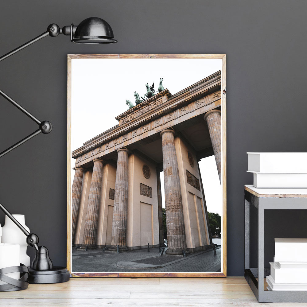 Poster vom Brandenburger Tor. Bild im hellen Holzbilderrahmen gerahmt und an einer grauen Wand stehend.