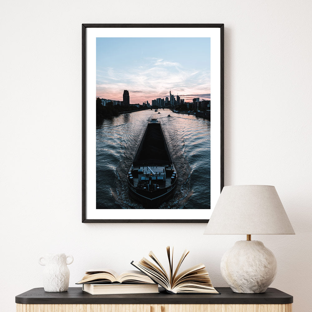 Frankfurt am Main Poster. Boot fährt über den Main, im Hintergrund die Frankfurter Skyline zum Sonnenuntergang. Bild mit weißen umlaufenden Rand. Gerahmt im schwarzen Rahmen über einer Kommode.