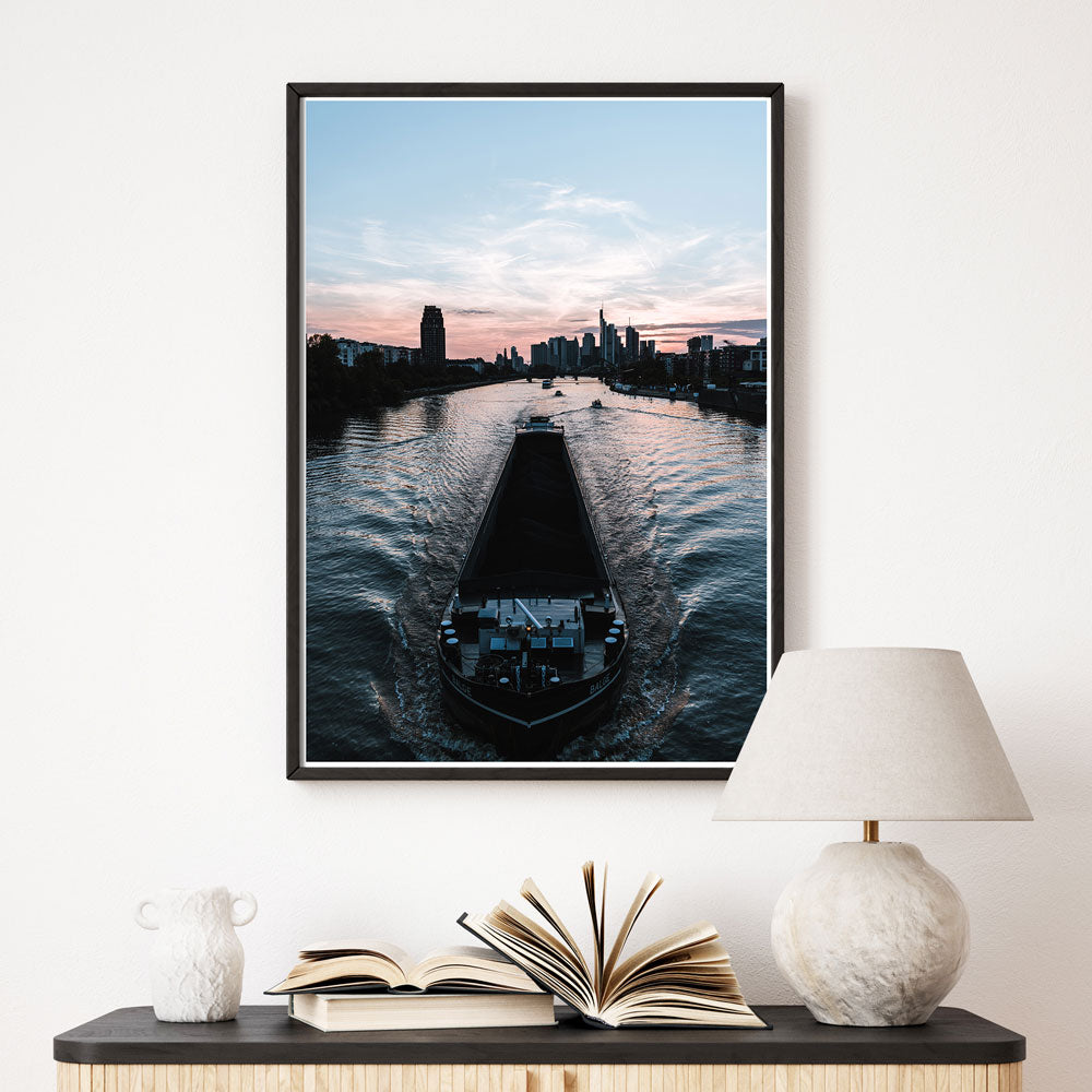 Frankfurt am Main Poster. Boot fährt über den Main, im Hintergrund die Frankfurter Skyline zum Sonnenuntergang. Bild im schwarzen Rahmen über einer Kommode.