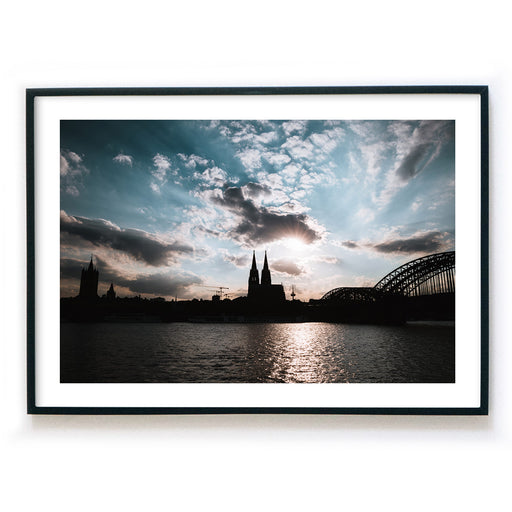 Köln Skyline Poster im Querformat. Sonnenuntergang über dem Kölner Dom bei leicht bewölkten blauen Himmel. Bild mit weißen Rand im Bilderrahmen.