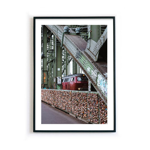 Bild mit Köln Motiv im schwarzen Bilderrahmen. Bahn fährt über die Hohenzollernbrücke, im Vordergrund sind die Liebesschlösser. Poster im Industrie Look und weißen Rand.