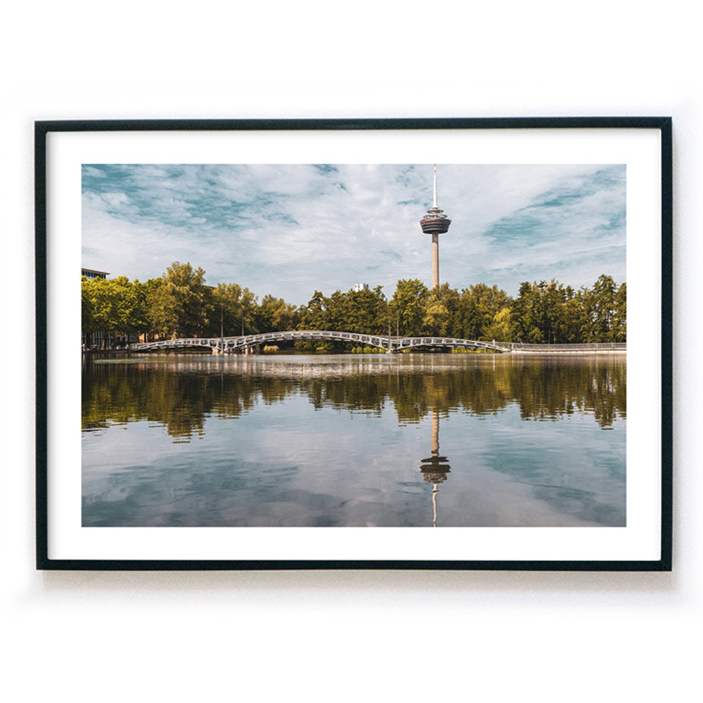Köln Bild im Bilderrahmen als Querformat. Motiv ist der Fernsehturm, eine Brücke und Bäume, die sich im See spiegeln. Bild mit weißen Rand.