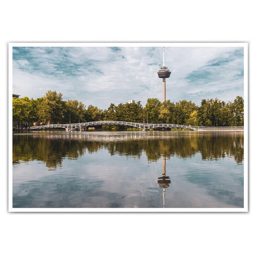 Köln Bild im Querformat. Motiv ist der Fernsehturm, eine Brücke und Bäume, die sich im See spiegeln. Sommertag mit blauem Himmel.