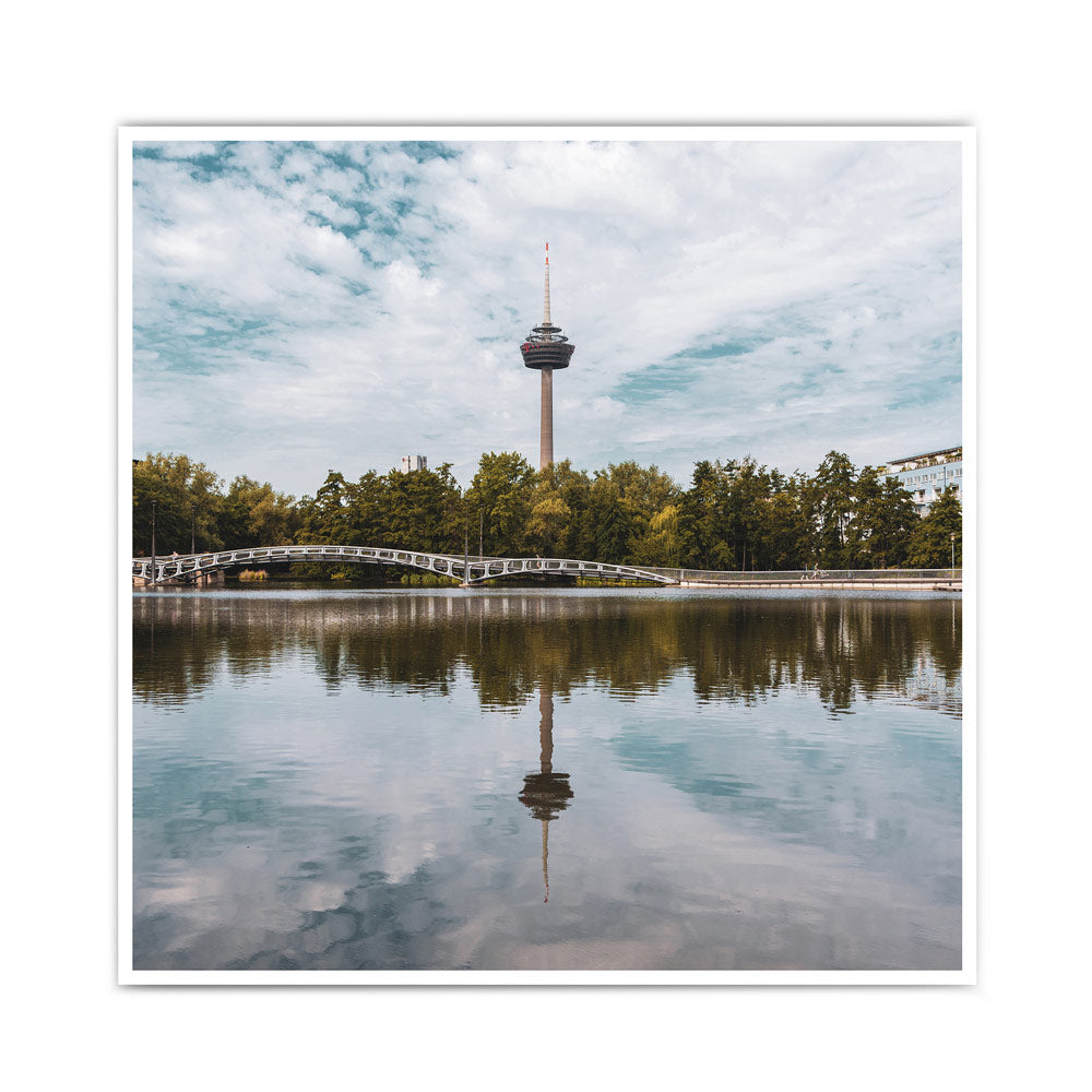 Quadratisches Sommer Köln Bild. Fernsehturm ist mittig im Bild und spiegelt sich im See im Vordergrund.