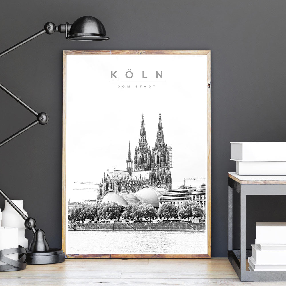 Köln Bild in einem Holzrahmen angelehnt an eine graue Wand. Motiv vom Rhein und dem Kölner Dom. Filter in schwarz-weiß und lässt das Bild wie gezeichnet wirken. Schriftzug im Bild - Köln Dom Stadt.