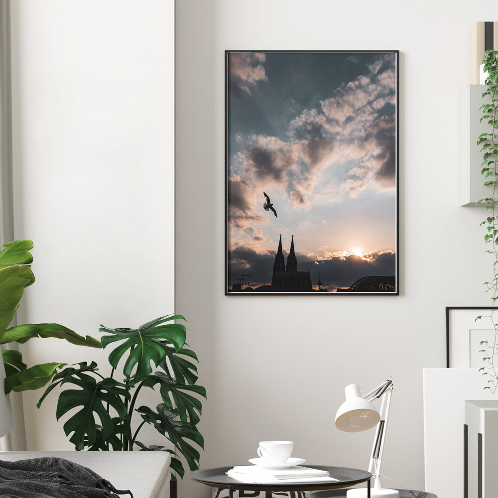 Köln Poster vom Sonnenuntergang über dem Kölner Dom. Über dem Dom fliegt eine Möwe. Kraftvoller blauer Himmel mit Wolken und Sonnenstrahlen. Aufgehangen im schwarzen Rahmen an der Wand im Wohnzimmer.